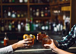 Three people clinking glasses, possibly in a bar or pub setting, with shelves of bottles visible in the background.