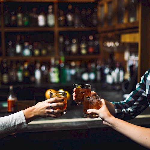Three people clinking glasses, possibly in a bar or pub setting, with shelves of bottles visible in the background.