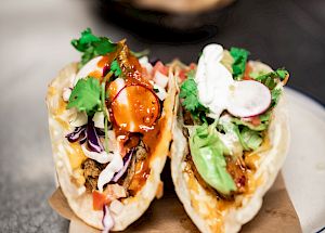 Two tacos filled with various ingredients including greens, radish slices, and a creamy sauce placed on a plate lined with brown paper.