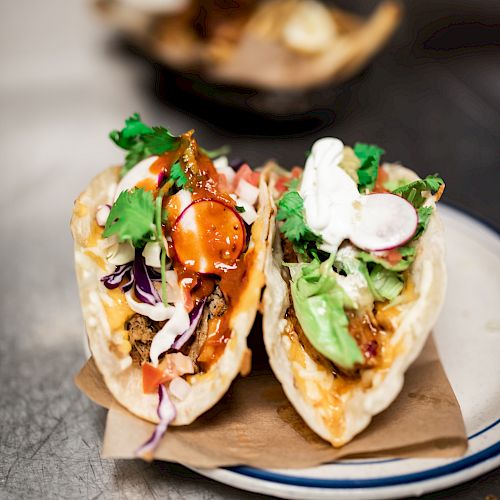 Two tacos filled with various ingredients including greens, radish slices, and a creamy sauce placed on a plate lined with brown paper.