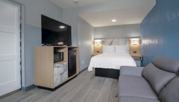 A modern hotel room with a bed, television, mini-fridge, sofa, and stylish decor in gray and white tones.