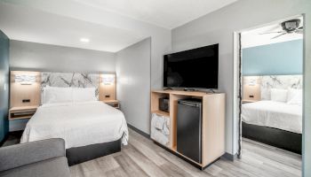 A modern hotel room with a bed, TV, mini-fridge, and sofa on wooden flooring. A mirror reflects part of the room's decor.