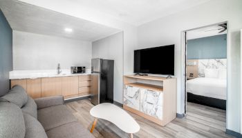 A modern living area with a grey couch, coffee table, kitchenette, TV on a cabinet, and a door leading to a bedroom.