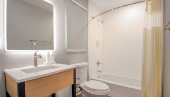 This image shows a modern bathroom featuring a sink with a vanity, a mirror, a toilet, and a bathtub with a shower curtain, all under bright lighting.