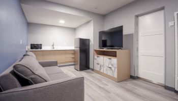 A modern living room with gray sofa, kitchenette with appliances, TV on a cabinet, wooden floor, and blue and white walls, next to a white door.