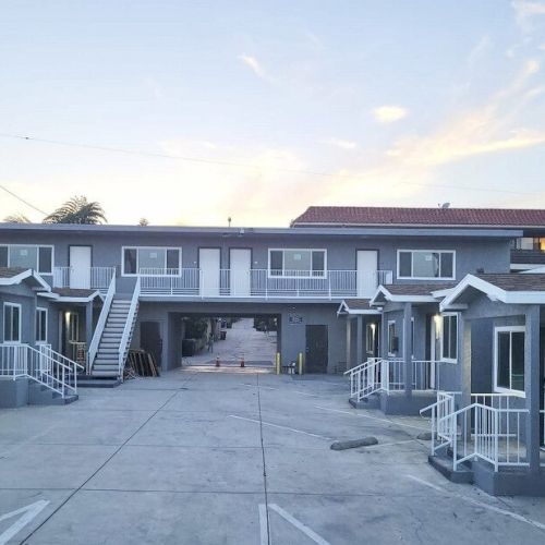 The image shows a small motel or apartment complex with multiple units, two levels, and a central staircase, with a clear sky in the background.