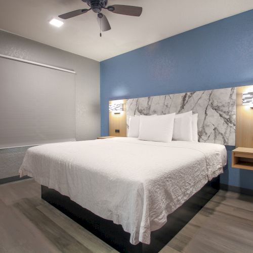 A modern hotel room with a double bed, white linens, blue accent wall, ceiling fan, and bedside lamps.