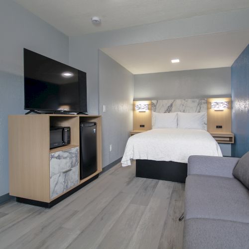 A modern hotel room with a bed, TV on a stand, mini fridge, and a grey sofa on a wooden floor.