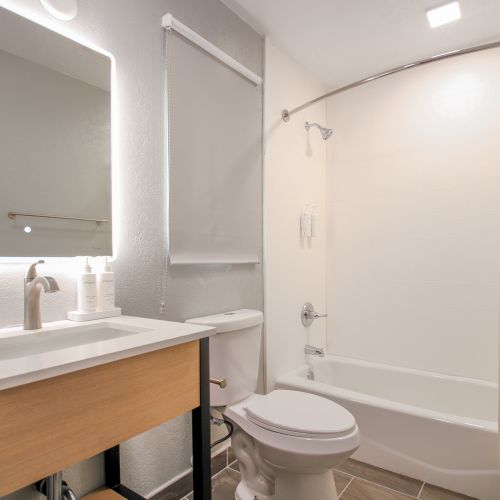 The image shows a modern bathroom with a lit vanity mirror, a wooden vanity sink, a toilet, and a shower area with a white curtain.