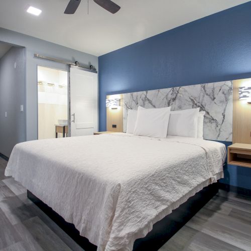 A modern hotel room with a king-sized bed, blue accent wall, and a view into a brightly lit bathroom through a sliding door, is shown.