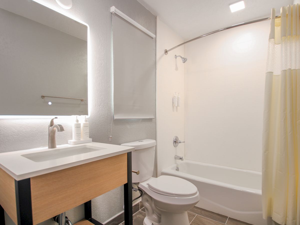 The image shows a modern bathroom with a lit vanity mirror, sink, toilet, bathtub-shower combination, and a yellow-and-white shower curtain, ending the sentence.