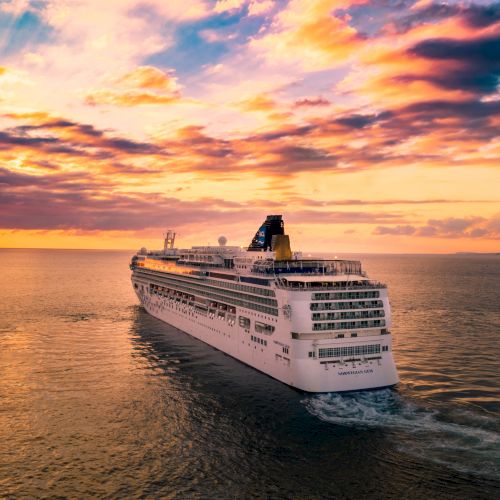 A cruise ship sails on calm waters, with a beautiful sunset and colorful clouds in the background, creating a serene and picturesque scene.