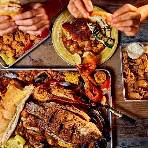 This image shows a seafood feast with grilled fish, shrimp, corn, lemon wedges, and bread rolls, being enjoyed by two people at a wooden table.