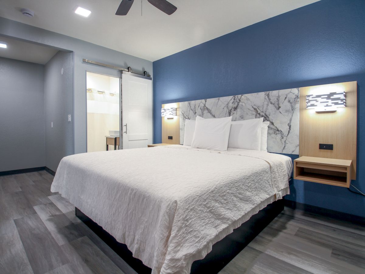 A modern hotel room with a large bed, blue accent wall, wall-mounted lighting, and a sliding barn door leading to the bathroom.