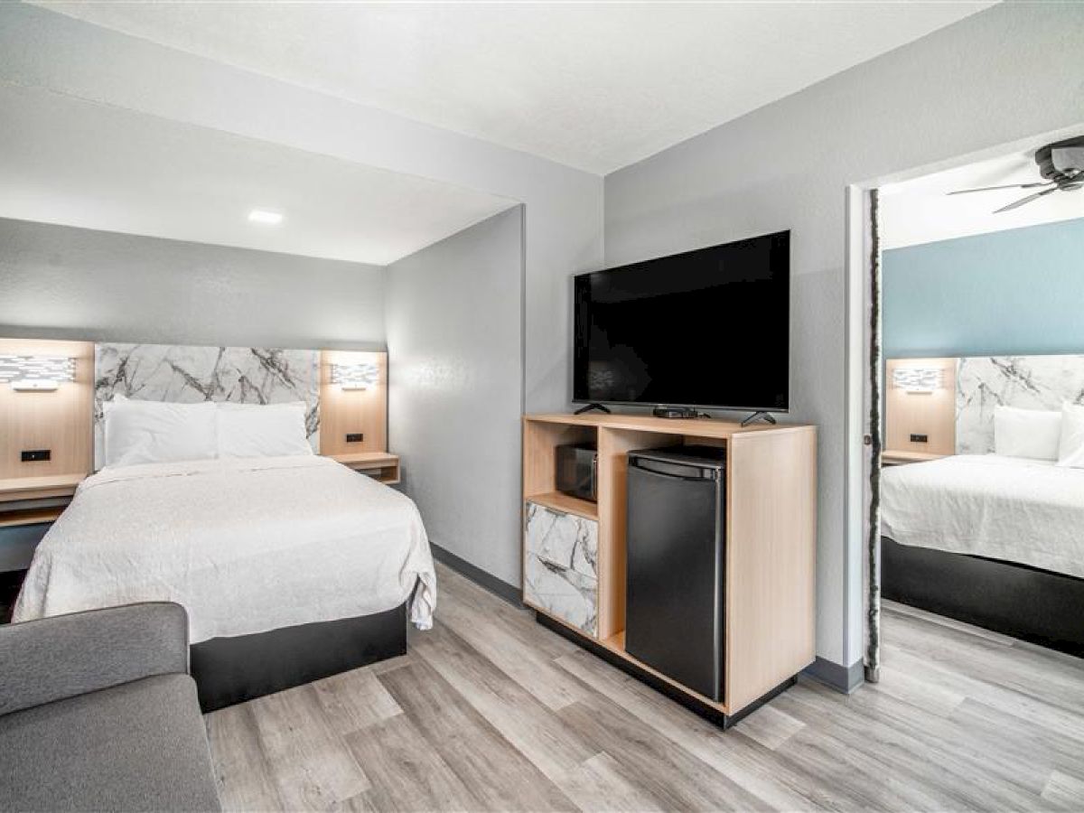 The image shows a modern hotel room with a bed, wall-mounted TV, mini fridge, and wooden flooring, reflecting in a mirror.