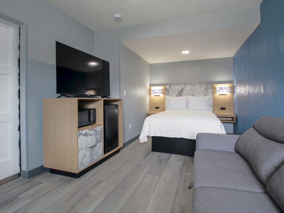 This image shows a modern hotel room with a bed, TV, mini-fridge, and a gray sofa in a minimalist design.