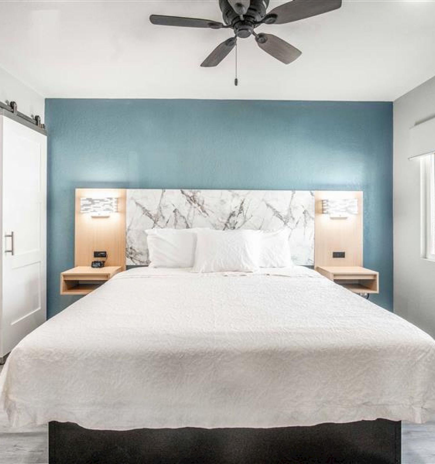 A modern bedroom with a double bed, blue accent wall, marble headboard, ceiling fan, and a sliding door leading to a bathroom.