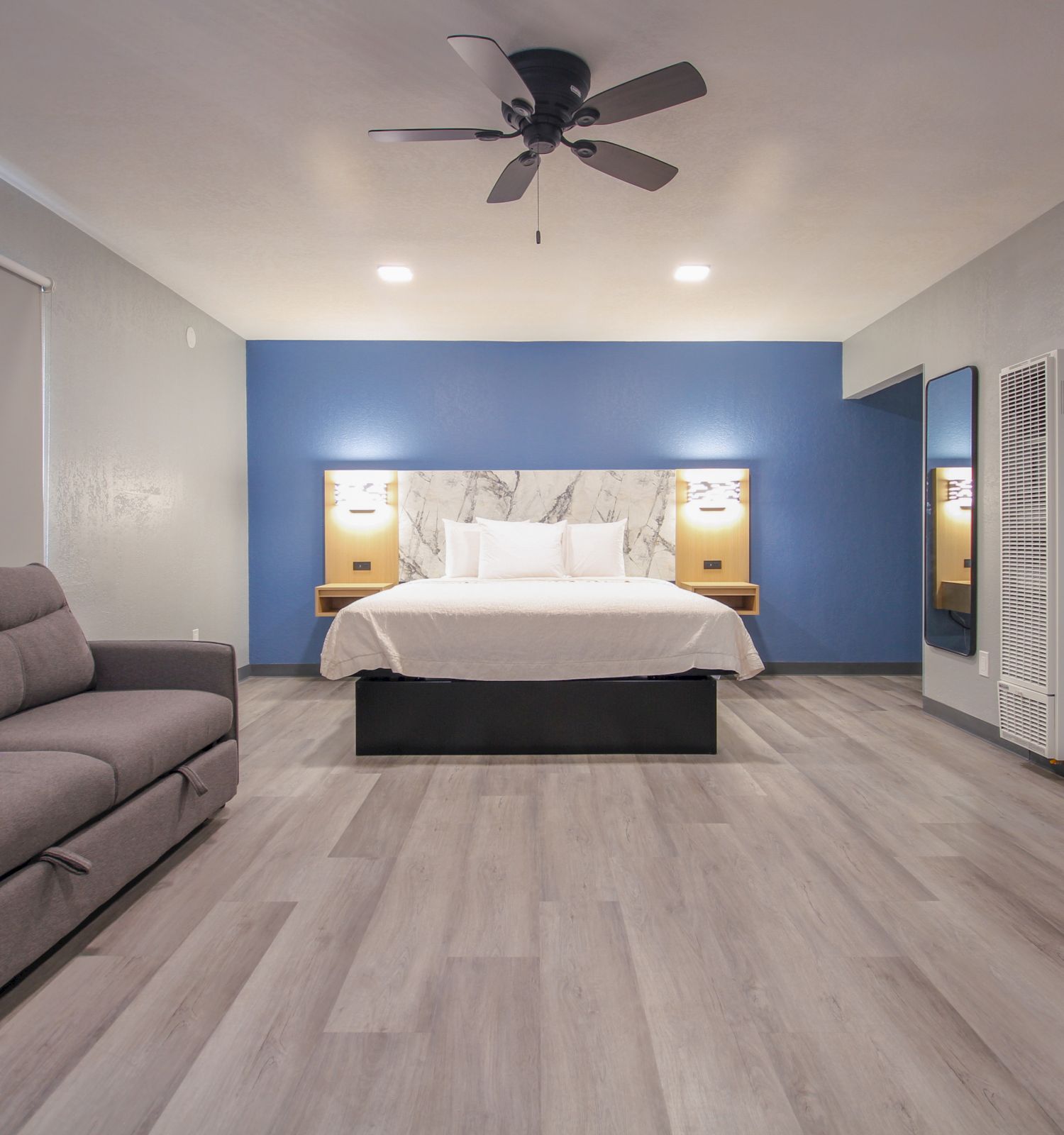 The image shows a modern hotel room with a large bed, blue accent wall, sofa, ceiling fan, wooden flooring, nightstands, and lamps on either side of the bed.