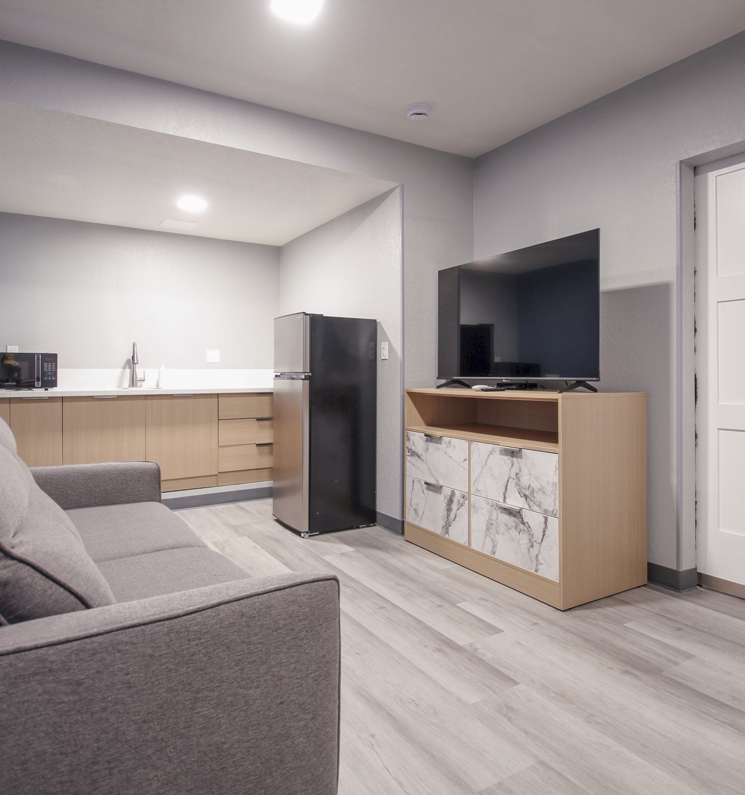 A modern living area with a gray sofa, TV on a stand, compact kitchen with a fridge and microwave, blue wall, and light wooden flooring.