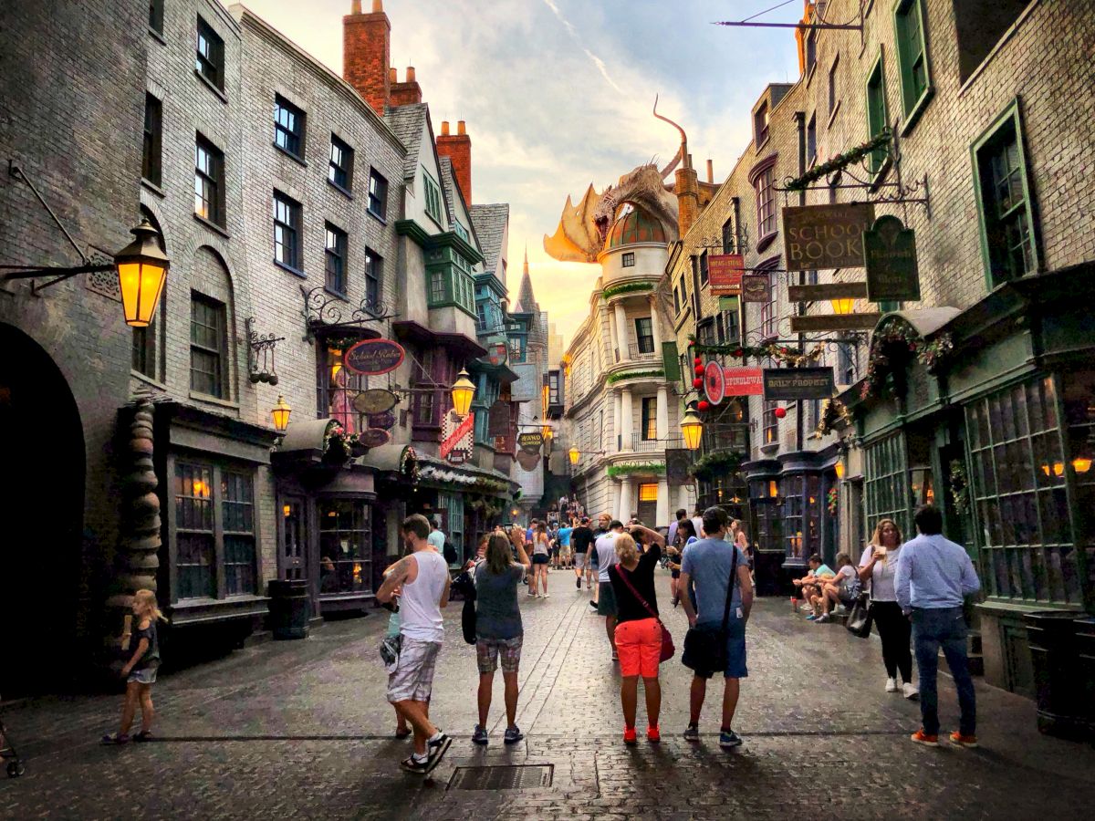 A lively cobblestone street scene featuring various shops, lit lamps, and a dragon perched atop a building in the background under a twilight sky.