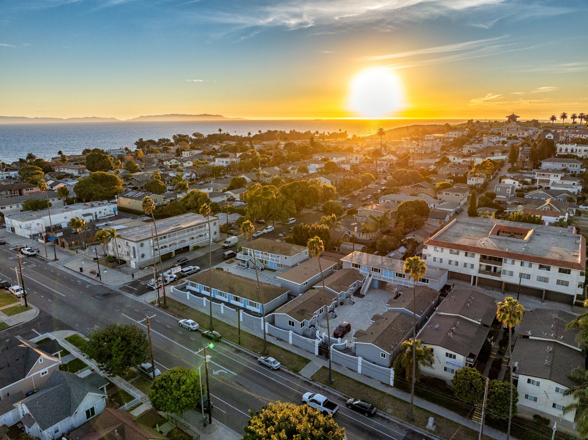 The image shows a seaside neighborhood at sunset, with palm trees, residential buildings, and a view of the ocean in the background.