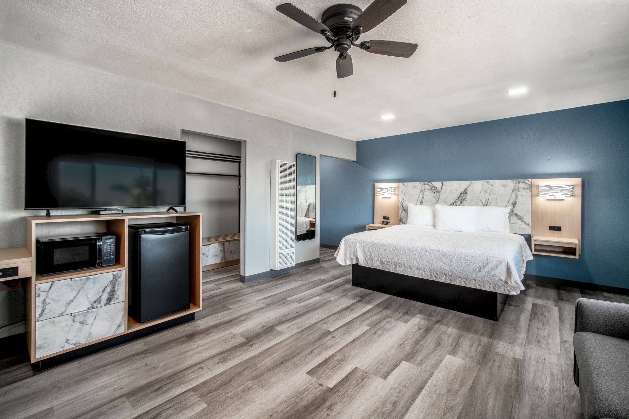 A modern hotel room with a bed, TV, ceiling fan, and kitchenette. Blue accent wall, wood flooring, and minimalistic decor complete the look.