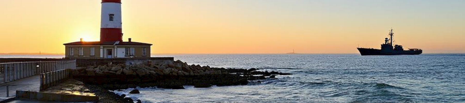 A lighthouse stands by the sea at sunset with a ship in the distance, waves crashing against the rocky shore.