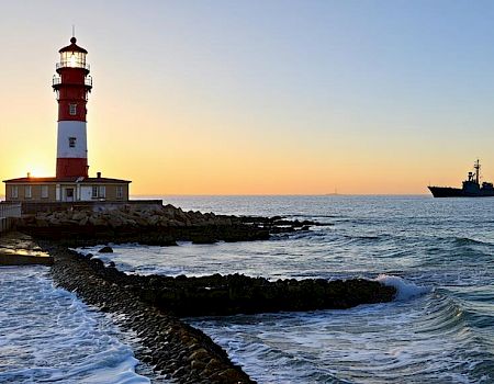 A lighthouse stands by the sea at sunset with a ship in the distance, waves crashing against the rocky shore.