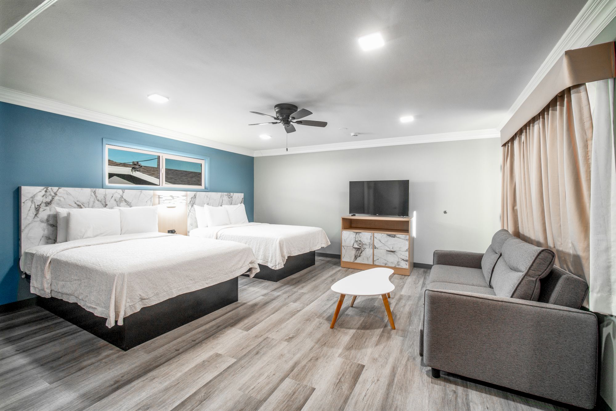 The image shows a hotel room with two beds, a couch, TV, coffee table, and artwork on the wall, featuring modern decor.