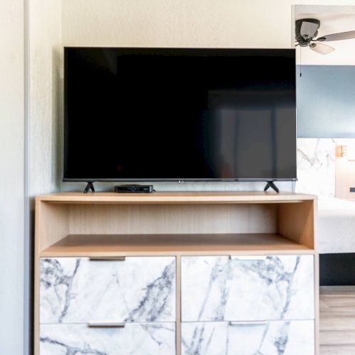 A flat-screen TV on a wooden stand with marble-patterned drawers, set in a room with a visible bed and ceiling fan.