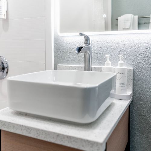 The image shows a modern bathroom sink with a square basin, a chrome faucet, and soap dispensers on a countertop, next to a large mirror.