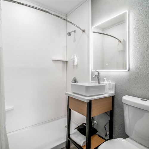 A modern bathroom with a shower, illuminated mirror, sink on a small table, and a toilet, featuring white and light gray tones.