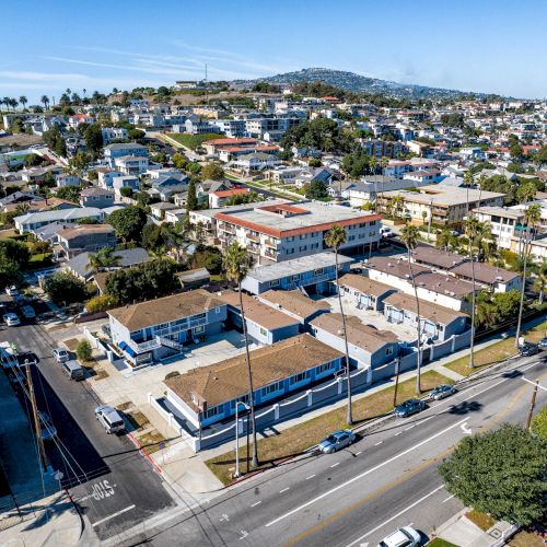 An aerial view of a suburban neighborhood with houses, trees, and a few cars on the road in a hilly area under clear blue skies.