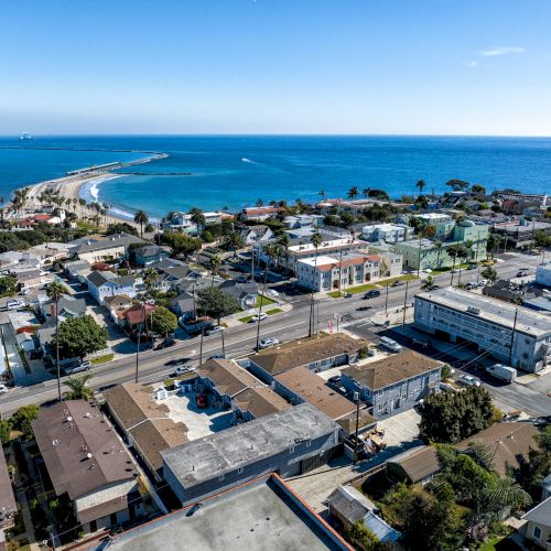 Aerial view of a coastal town with buildings, streets, and palm trees near the ocean. The sea and sky are clear and blue.