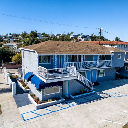 The image shows a two-story building with a brown roof, blue awnings, and white railings, surrounded by a parking area and other buildings.