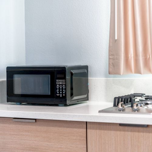 A kitchen counter features a black microwave and a gas stove, with light-colored cabinets and curtains in the background.