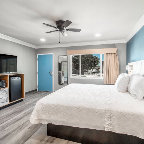 The image shows a modern hotel room with a bed, TV, cabinets, and a window. There's a ceiling fan and a light-colored floor, creating a simple look.