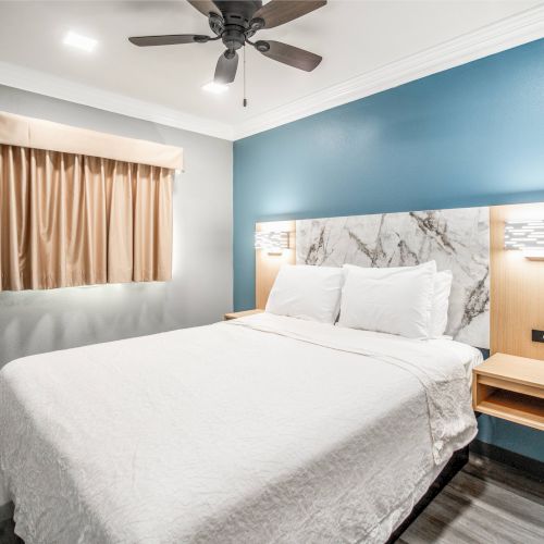 A modern bedroom with a white bed, blue accent wall, ceiling fan, small window, and two wall lights on each side of the bed.