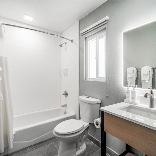 A modern bathroom with a shower, toilet, vanity, and illuminated mirror. Towels hang by the shower; a window provides natural light.