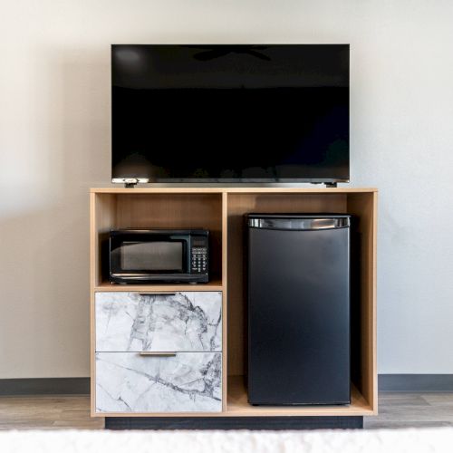 A TV is on a wooden stand with a microwave, mini-fridge, and a drawer. The setup is placed against a plain wall.