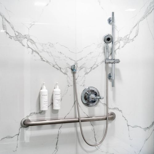 The image shows a marble-tiled shower with a showerhead, grab bar, and two bottles of shampoo or body wash on a shelf.