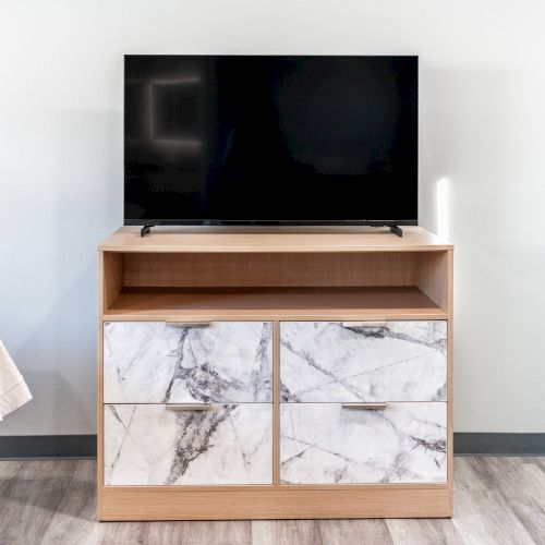 A flat-screen TV sits on a wooden cabinet with marble-patterned drawers in a room with a bed partially visible on the left side.