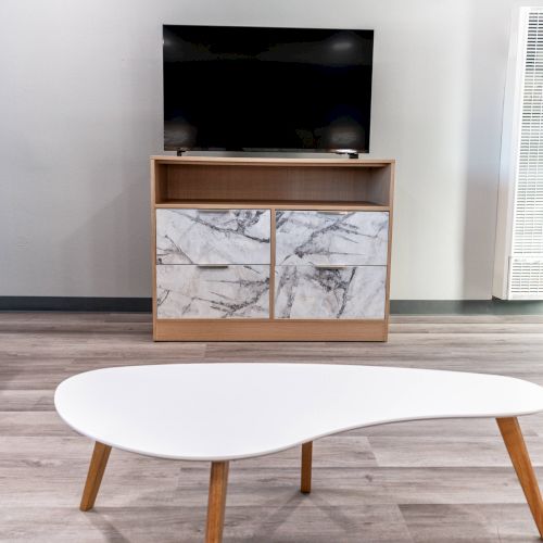 A minimalist living room with a TV on a wooden stand, a gray sofa, and a white, uniquely shaped coffee table on a wooden floor.