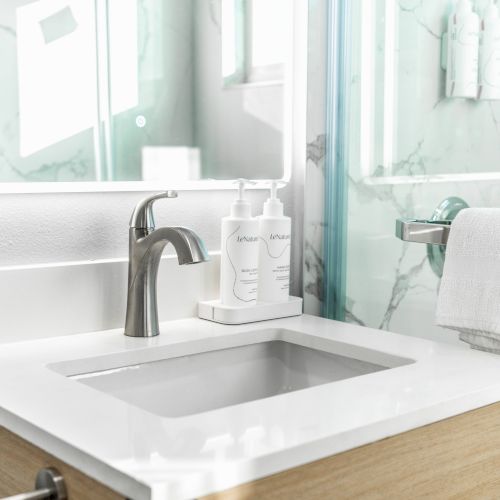 A modern bathroom sink with a chrome faucet, two bottles on the counter, and a towel on the glass shower door handle.