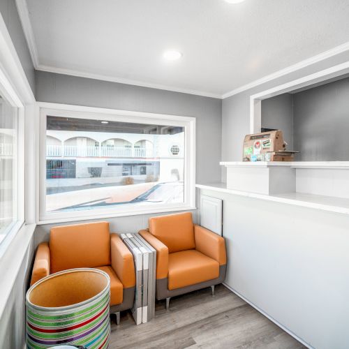 A small waiting area with two orange chairs and a stack of colorful rings by the window, featuring a reception counter and bright lighting.