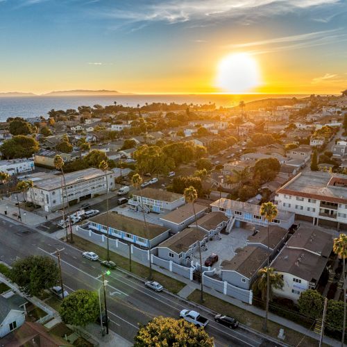 Aerial view of a coastal town at sunset, with ocean in the background, residential buildings, and tree-lined streets.