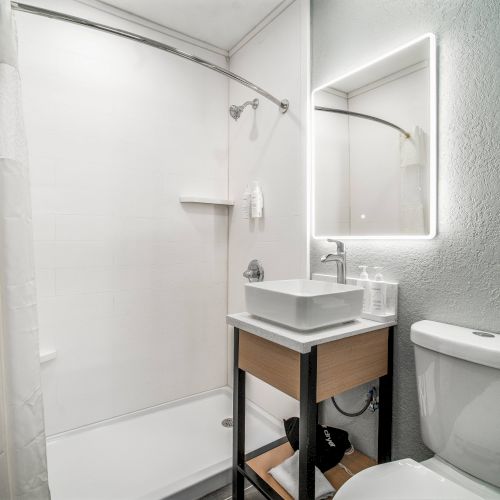 A modern bathroom with a shower, illuminated mirror, sink, and toilet. White towels hang beside the shower curtain.