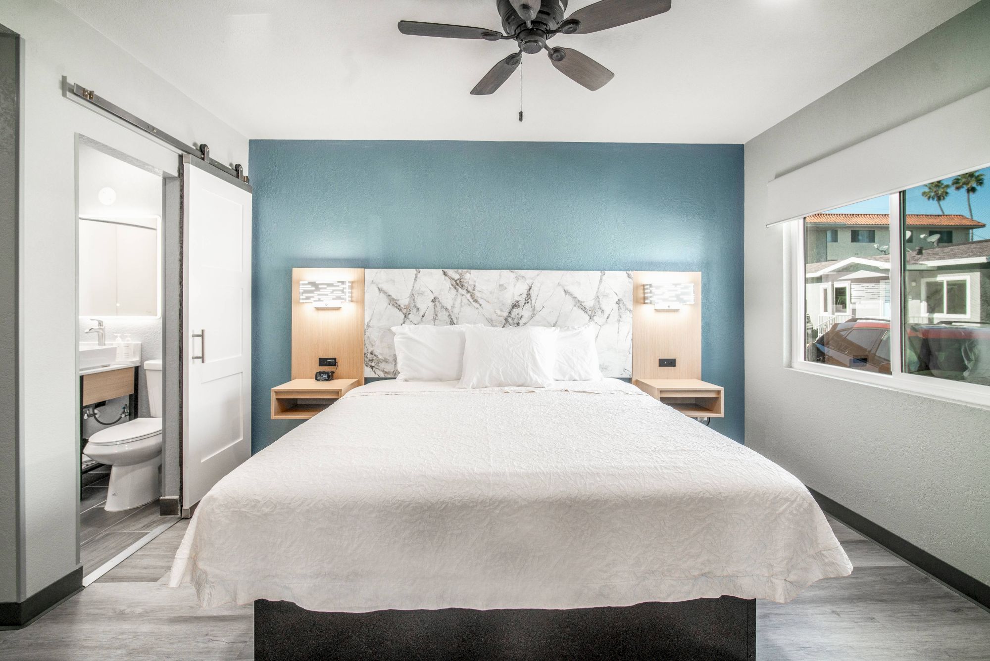 A modern bedroom with a queen bed, marble headboard, ceiling fan, sliding bathroom door, and a window with an outdoor view completes the setup.