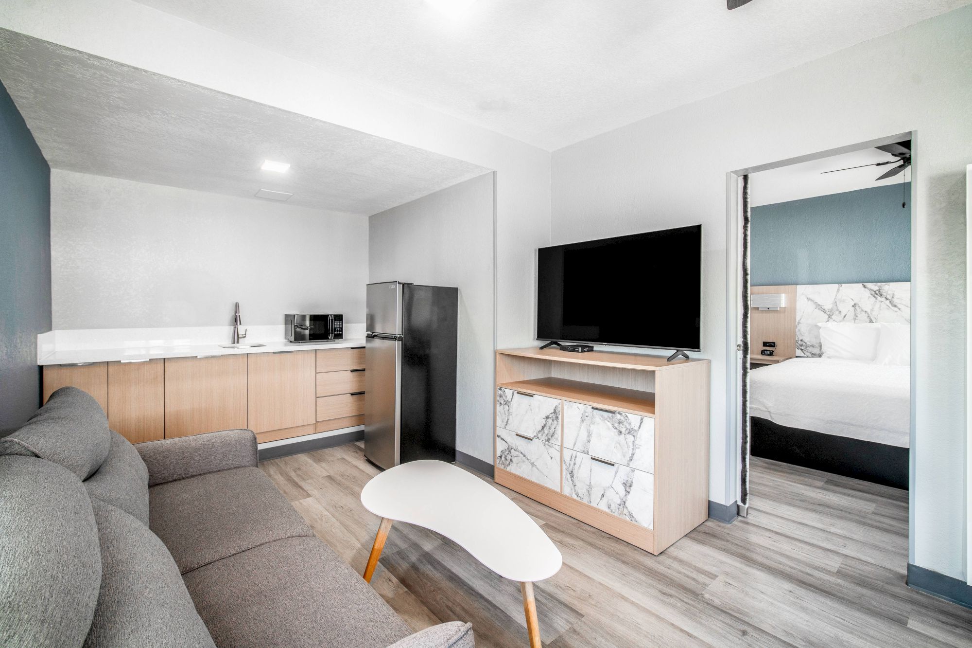 A modern living area with a sofa, table, TV, kitchenette, and a doorway leading to a bedroom with a bed.