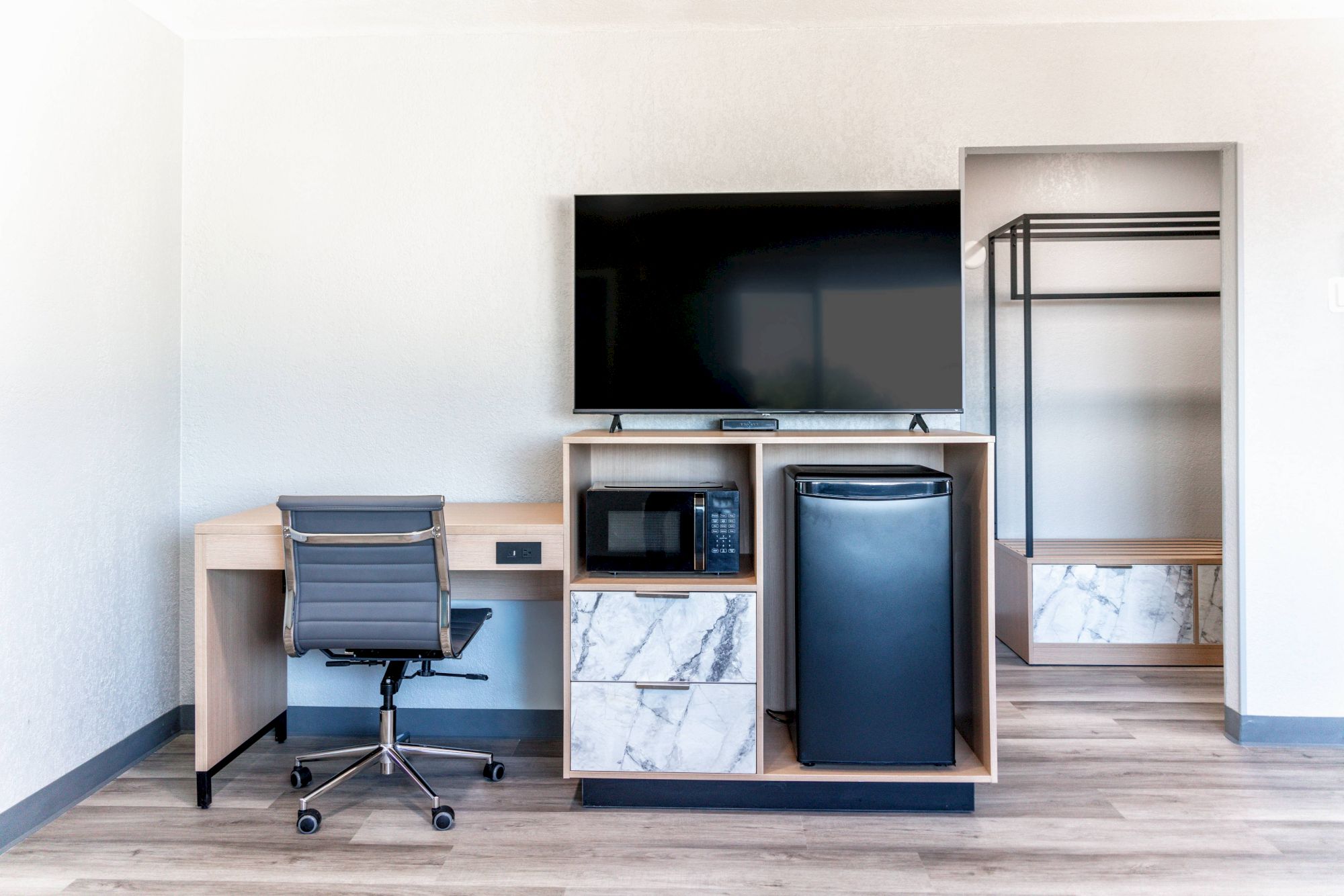 A modern room setup with a desk, chair, large TV, microwave, fridge, and open closet on a wooden floor.